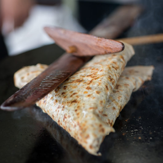 Folded crepe on a hot griddle being pressed with a wooden crepe trowel and scraper.
